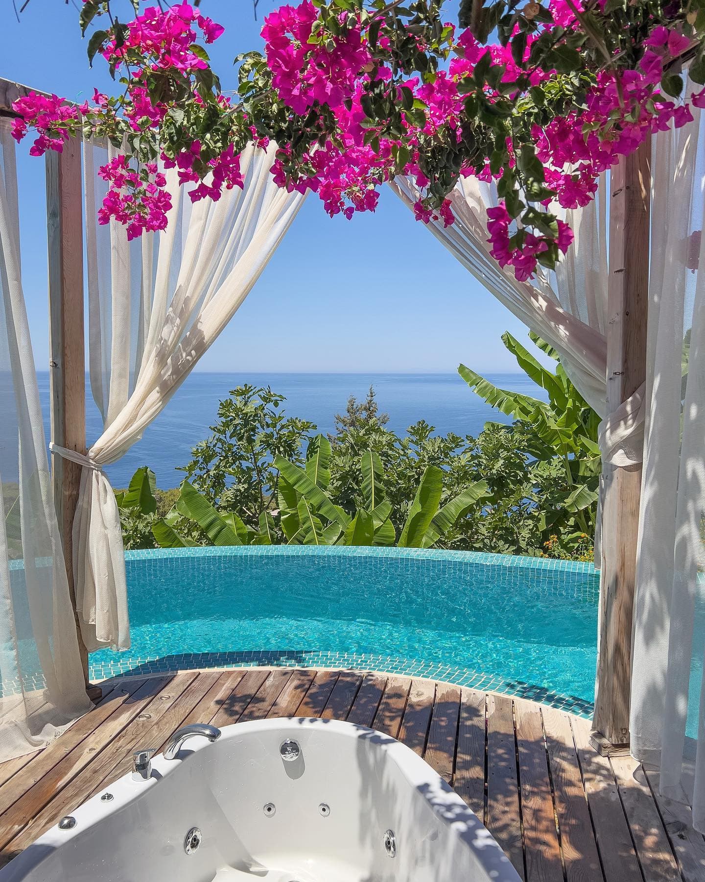 Infinity-Pool, Bananenpflanzen und Meer zwischen magentafarbener Bougainvillea und Vorhängen.