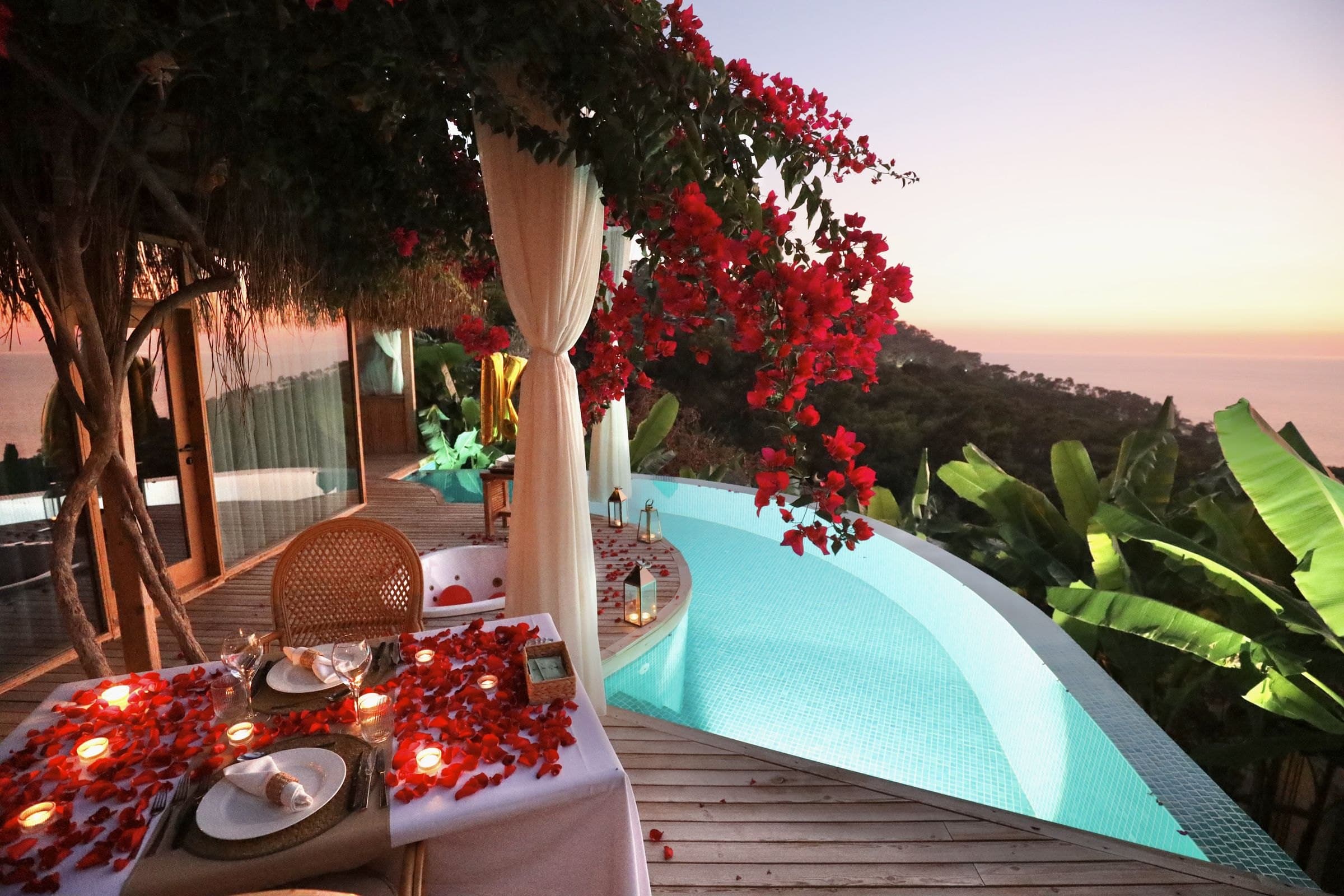 Ein Tisch mit Kerzen und Rosenblättern unter Bougainvillea in der Abenddämmerung, Infinity-Pool und Meer.