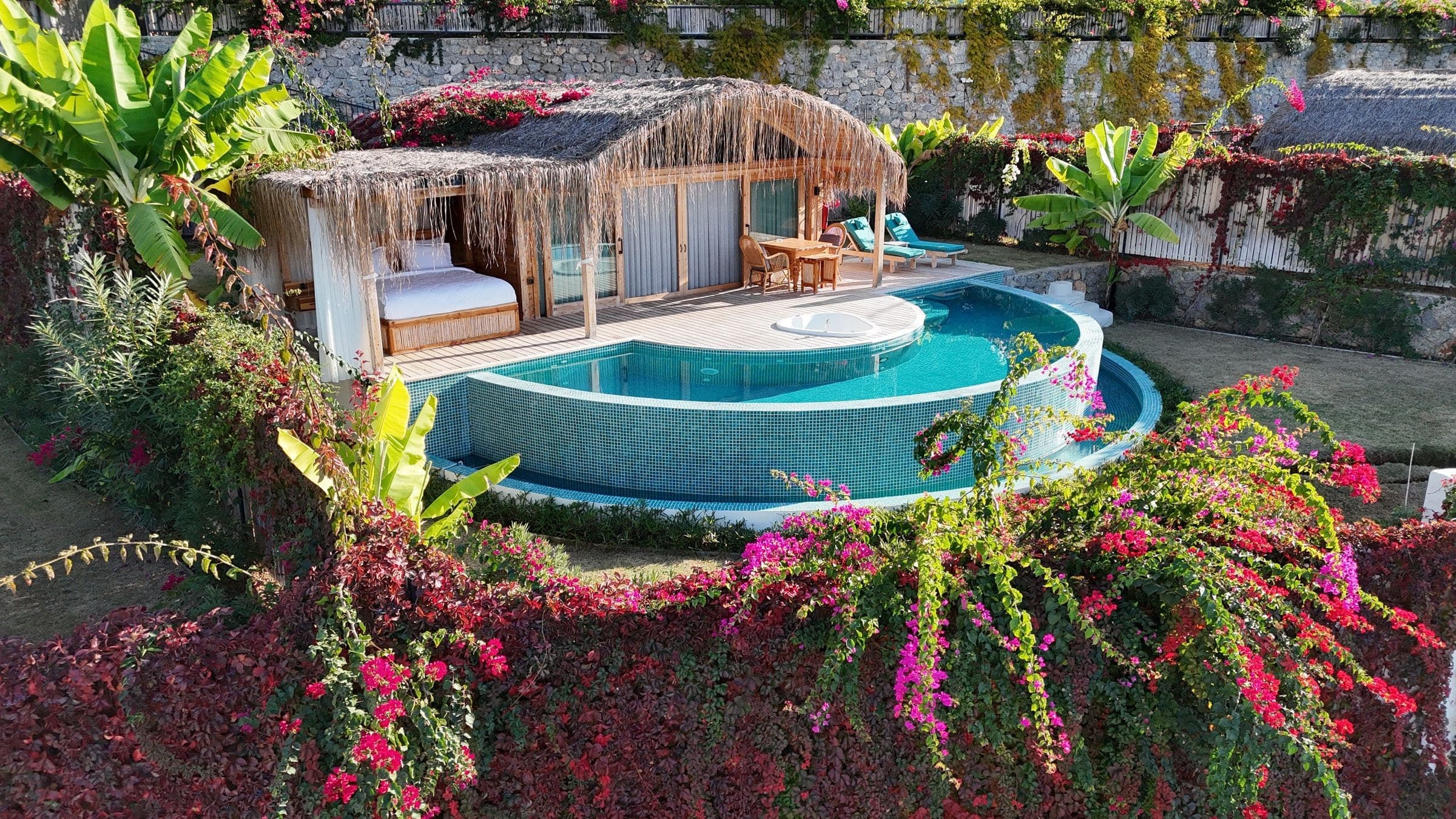 Çınar aus niedriger Drohnenhöhe: vorn eine Hecke aus magentafarbener und roter Bougainvillea, dahinter halbmondförmiger Pool, Whirlpool-Insel und Schilfhütte.